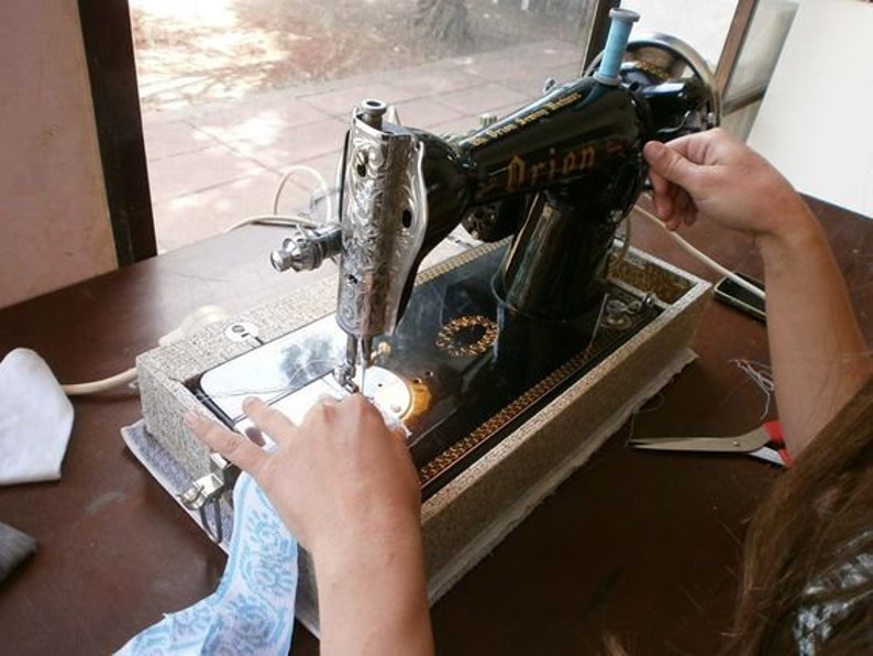 May include: A black sewing machine with a silver design and the word "Orion" on the side. The machine is on a wooden table with a white cloth and a pair of scissors.