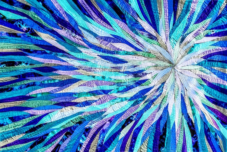 Op de afbeelding: Een kleurrijk abstract quilt met een uitbarsting van blauwe, turquoise, paarse en witte stofstroken die vanuit het midden naar buiten stralen.