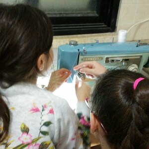 May include: Two people are working on a blue sewing machine. One person is threading the needle while the other person is holding the fabric.