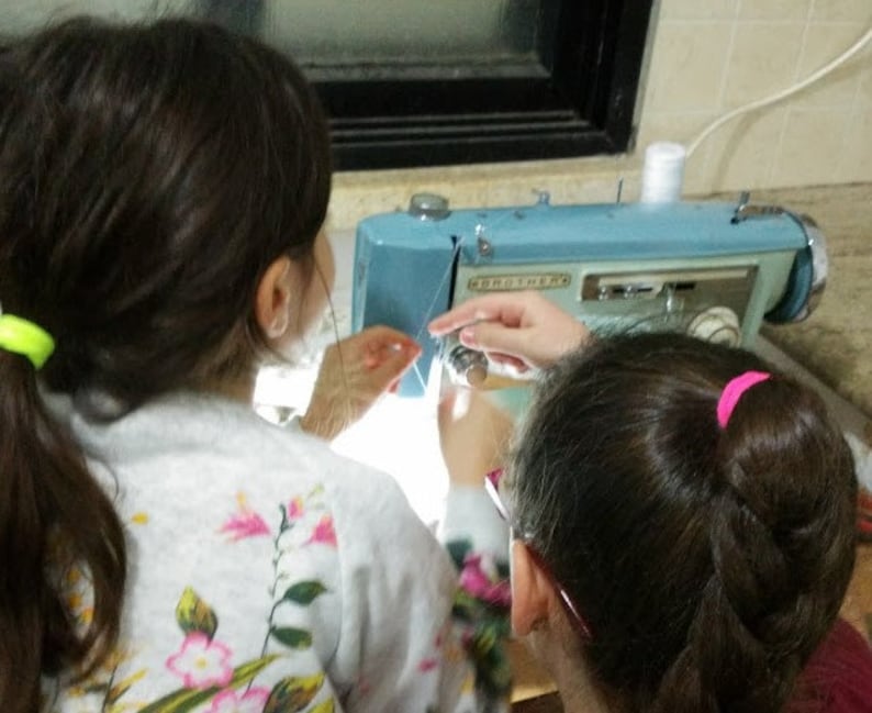 May include: Two people are working on a blue sewing machine. One person is threading the needle while the other person is holding the fabric.