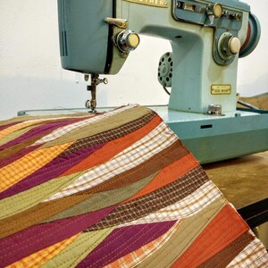 Może przedstawiać: Zabytkowa, jasnoniebieska maszyna do szycia Brother ze złotymi literami i pokrętłami. Kolorowy, pikowany kawałek materiału z ukośnymi paskami w odcieniach pomarańczu, fioletu, zieleni i brązu jest umieszczony pod igłą.