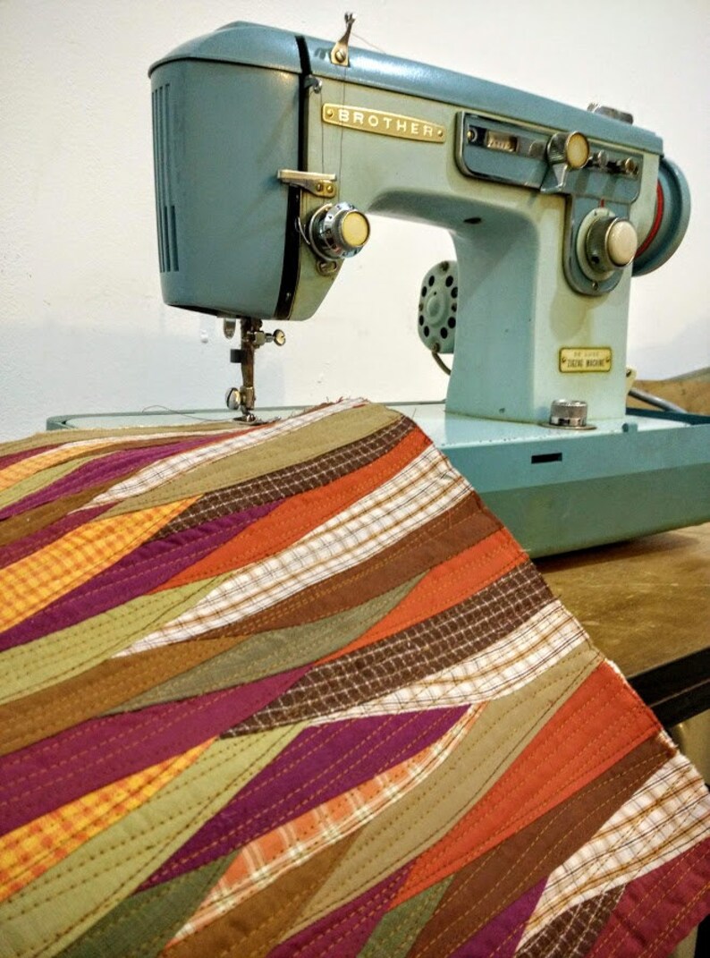 Może przedstawiać: Zabytkowa, jasnoniebieska maszyna do szycia Brother ze złotymi literami i pokrętłami. Kolorowy, pikowany kawałek materiału z ukośnymi paskami w odcieniach pomarańczu, fioletu, zieleni i brązu jest umieszczony pod igłą.