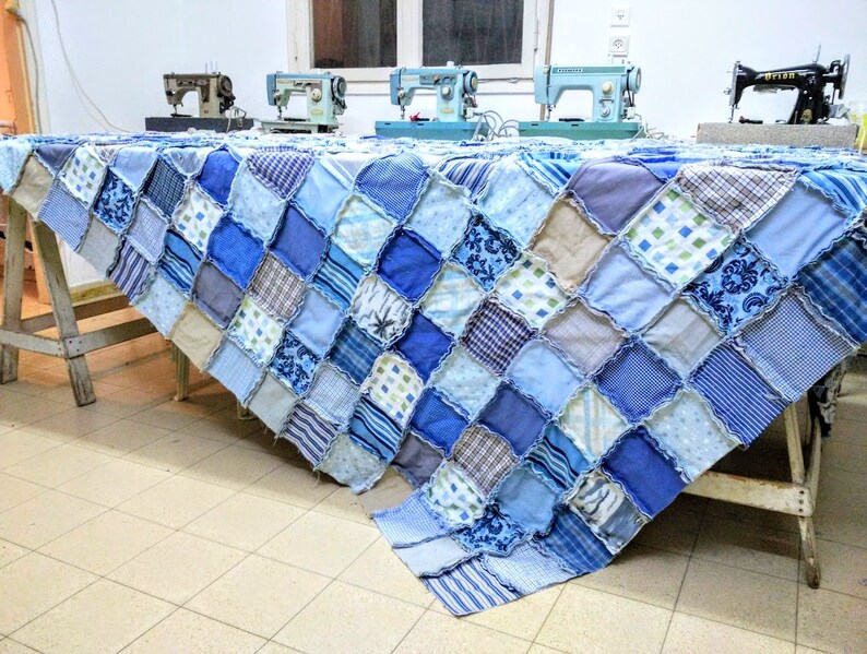 May include: A blue and beige patchwork quilt made from squares of different fabrics. The quilt is draped over a table with four sewing machines in the background.