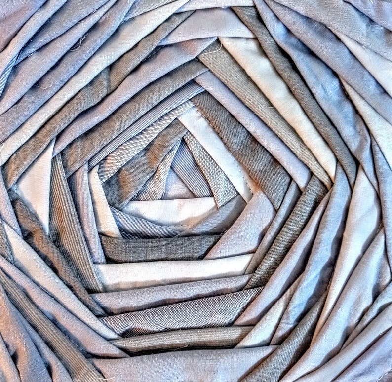 Pu&ograve; includere: Primo piano di una trapunta circolare realizzata con diverse tonalit&agrave; di tessuto grigio. Il tessuto &egrave; cucito a spirale, creando un design testurizzato e intricato.