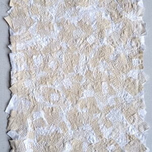 May include: A handmade textile art piece featuring a patchwork design. The piece is composed of various fabric squares in off-white and beige tones, stitched together. The texture is visible with visible stitching and varied fabric patterns.