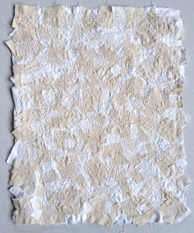 May include: A handmade textile art piece featuring a patchwork design. The piece is composed of various fabric squares in off-white and beige tones, stitched together. The texture is visible with visible stitching and varied fabric patterns.