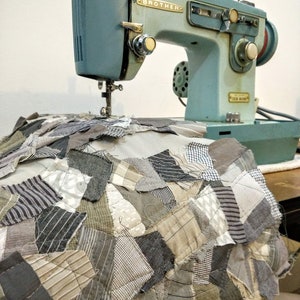 May include: A vintage Brother sewing machine in a light blue hue, positioned above a patchwork quilt. The quilt features a variety of fabric patterns in shades of gray, beige, and white. The machine's details are visible, including the needle and dials.