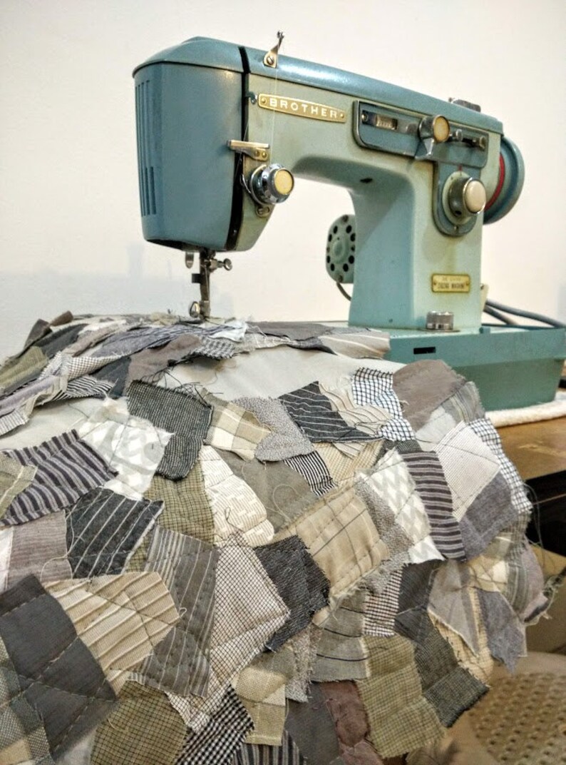 May include: A vintage Brother sewing machine in a light blue hue, positioned above a patchwork quilt. The quilt features a variety of fabric patterns in shades of gray, beige, and white. The machine's details are visible, including the needle and dials.