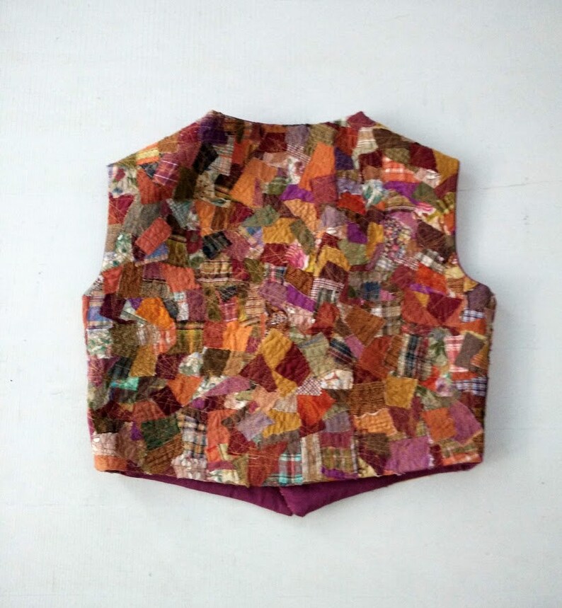Op de afbeelding: Een sleeveless vest gemaakt van een patchwork van verschillende stoffen in tinten bruin, oranje, rood en groen. Het vest heeft een hoge halslijn en een puntige onderkant.