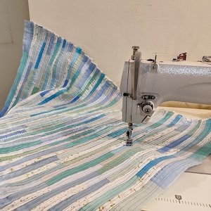 Puede incluir: Una m&aacute;quina de coser blanca con pedal est&aacute; cosiendo una tela de rayas azules y blancas. La tela est&aacute; hecha de m&uacute;ltiples tiras de diferentes tonos de azul y blanco.