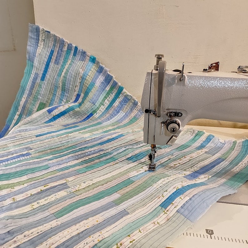 Puede incluir: Una m&aacute;quina de coser blanca con pedal est&aacute; cosiendo una tela de rayas azules y blancas. La tela est&aacute; hecha de m&uacute;ltiples tiras de diferentes tonos de azul y blanco.