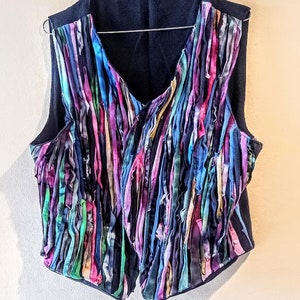 Puede incluir: Un chaleco sin mangas con base negra y una franja colorida hecha de tiras de tela en tonos azules, morados, rosas, verdes y amarillos.