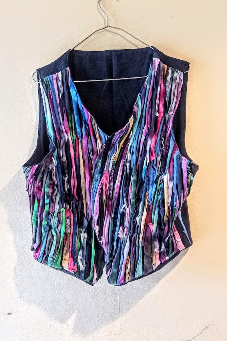 Puede incluir: Un chaleco sin mangas con base negra y una franja colorida hecha de tiras de tela en tonos azules, morados, rosas, verdes y amarillos.