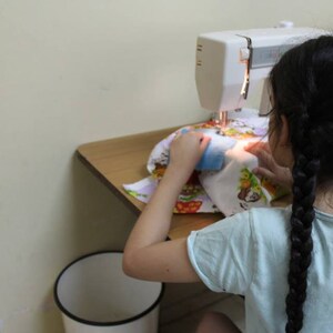 May include: A young person is sitting at a table using a white sewing machine to sew a colorful fabric with a cartoon design. The person is wearing a light blue shirt and has long black braids.