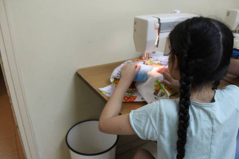 May include: A young person is sitting at a table using a white sewing machine to sew a colorful fabric with a cartoon design. The person is wearing a light blue shirt and has long black braids.