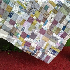May include: A patchwork quilt made from a variety of fabrics in shades of brown, gray, green, and purple. The quilt is made up of squares of different sizes and is hanging over a wooden surface.