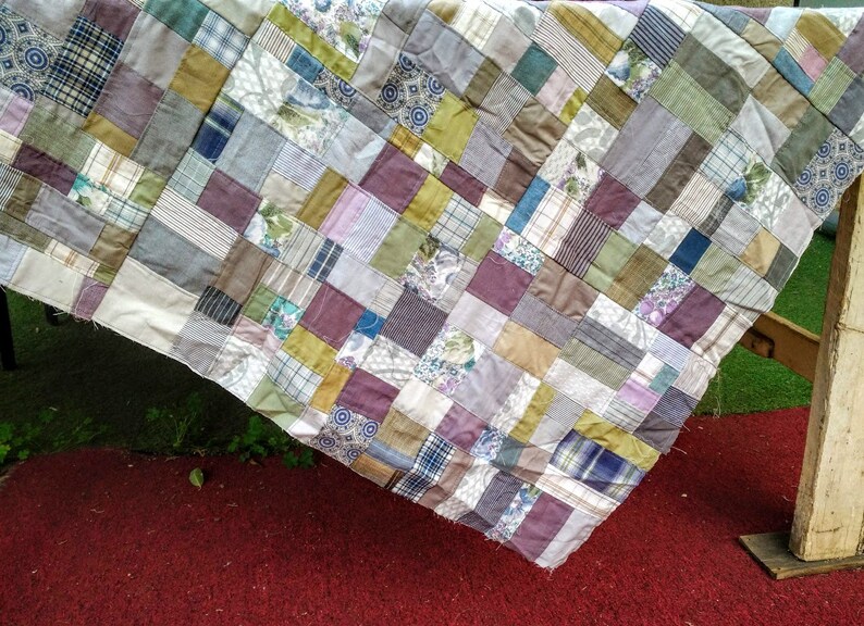 May include: A patchwork quilt made from a variety of fabrics in shades of brown, gray, green, and purple. The quilt is made up of squares of different sizes and is hanging over a wooden surface.
