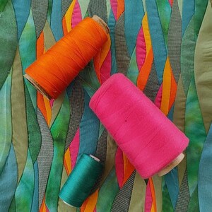 May include: Three spools of thread in different colours: orange, pink, and green. The spools are lying on a colourful fabric background with a wavy pattern.