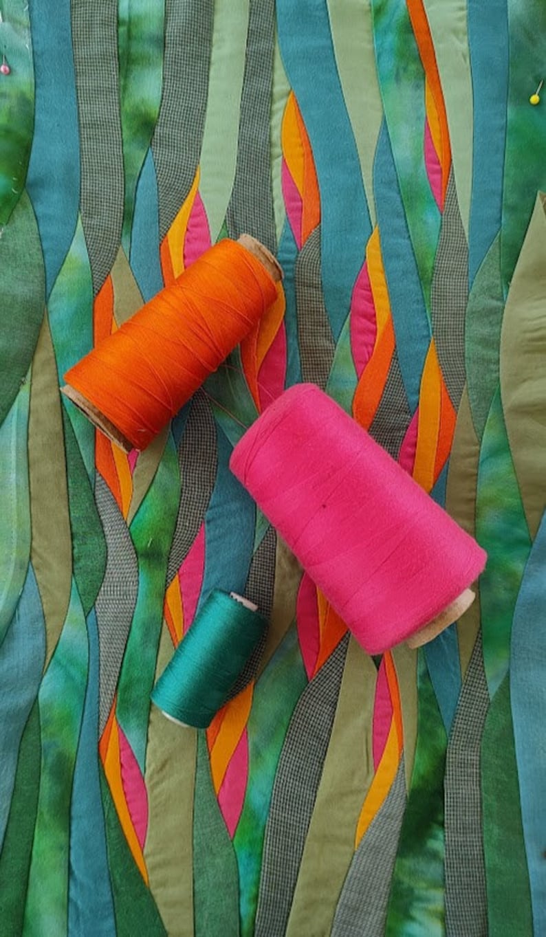 May include: Three spools of thread in different colours: orange, pink, and green. The spools are lying on a colourful fabric background with a wavy pattern.