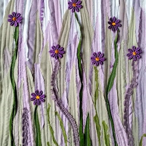 May include: A purple and green fabric art piece with a textured background and purple flowers. The flowers are made of fabric and have a yellow centre.