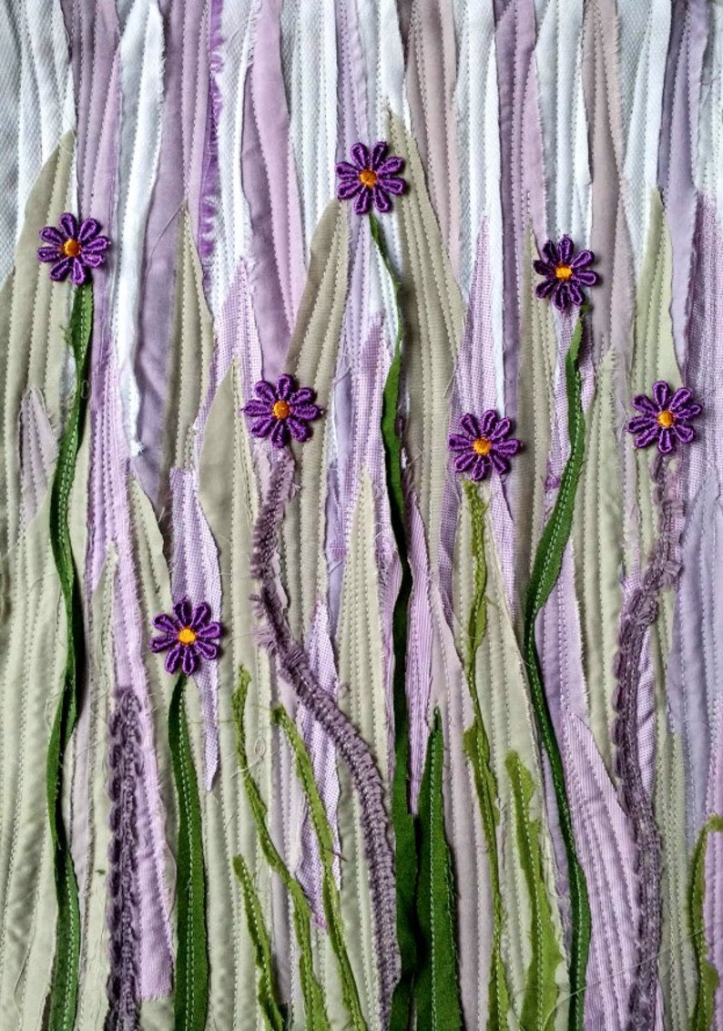 May include: A purple and green fabric art piece with a textured background and purple flowers. The flowers are made of fabric and have a yellow centre.