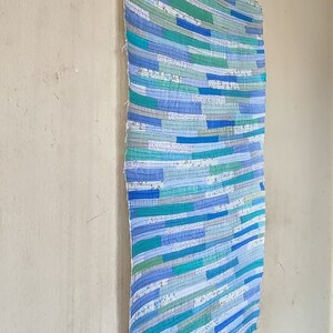 Puede incluir: Una colcha de retazos azul y verde con un patr&oacute;n de rayas. La colcha est&aacute; colgada en una pared.