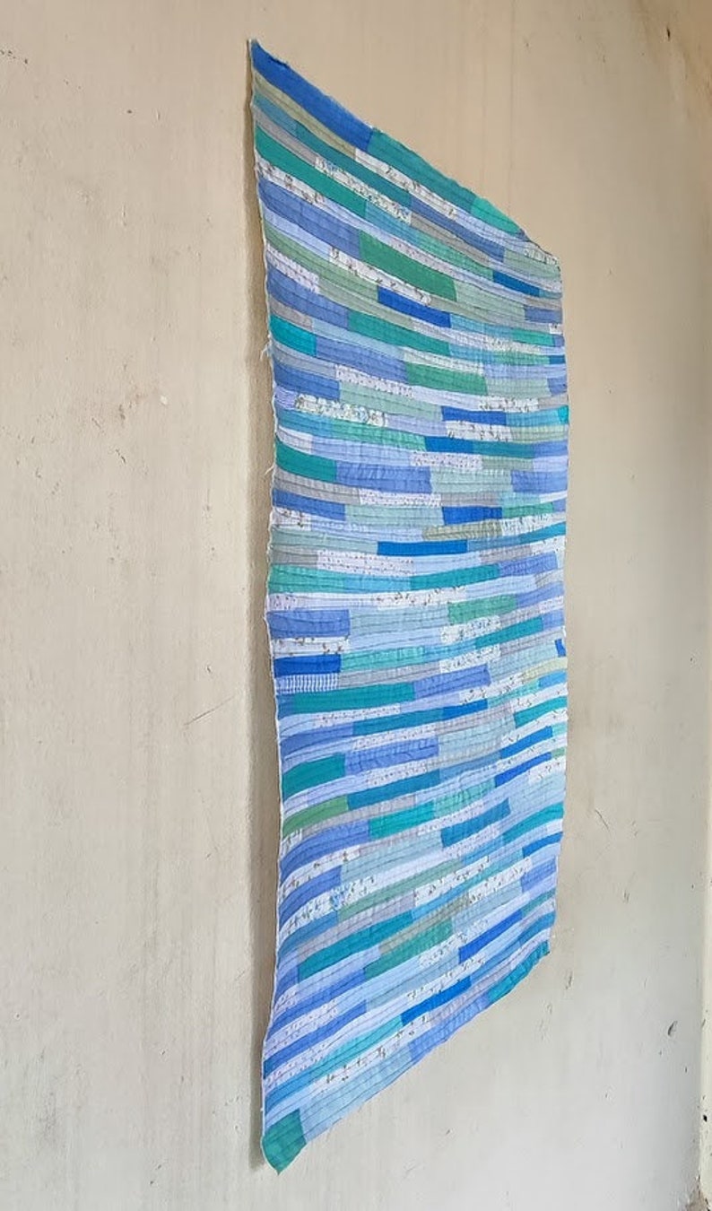 Puede incluir: Una colcha de retazos azul y verde con un patr&oacute;n de rayas. La colcha est&aacute; colgada en una pared.