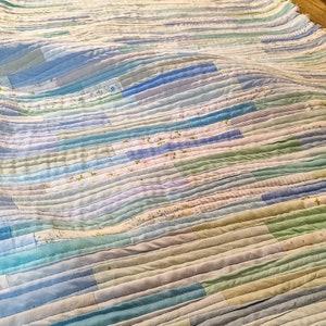 Puede incluir: Una colcha de retazos con rayas horizontales en tonos de azul, verde y blanco. La colcha est&aacute; hecha de varias telas con diferentes patrones, incluyendo estampados florales y colores s&oacute;lidos.