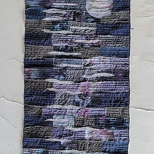 May include: A long, narrow wall hanging made of patchwork fabric in shades of blue, purple, and grey. The fabric is sewn together in a layered, abstract pattern, creating a textured and dimensional effect.