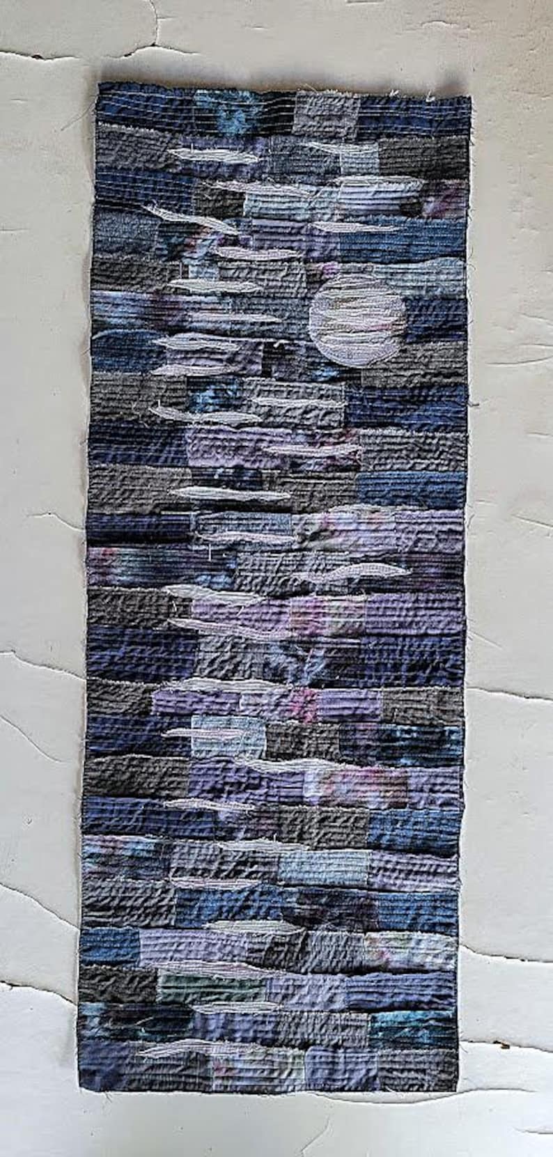 May include: A long, narrow wall hanging made of patchwork fabric in shades of blue, purple, and grey. The fabric is sewn together in a layered, abstract pattern, creating a textured and dimensional effect.