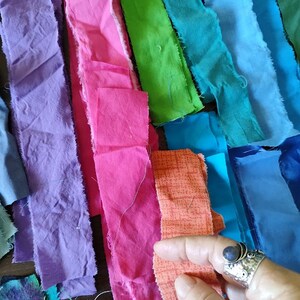 May include: Assortment of fabric scraps in various colours, including purple, pink, green, blue, and orange. The fabric scraps are arranged in a rainbow pattern and are lying on a wooden surface.