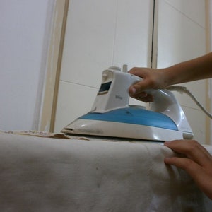 May include: A white and blue electric iron with a cord is being used to iron a white fabric on a table. A spray bottle with a red cap is visible in the background.