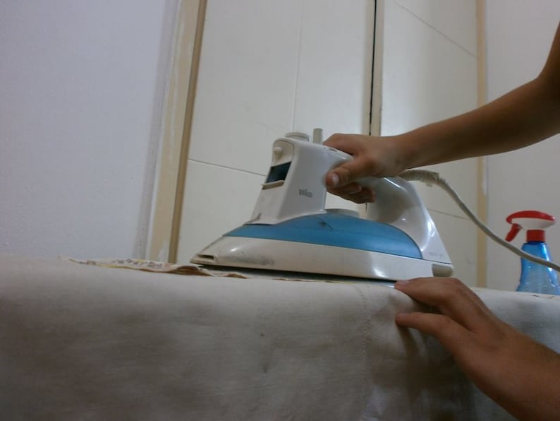 May include: A white and blue electric iron with a cord is being used to iron a white fabric on a table. A spray bottle with a red cap is visible in the background.