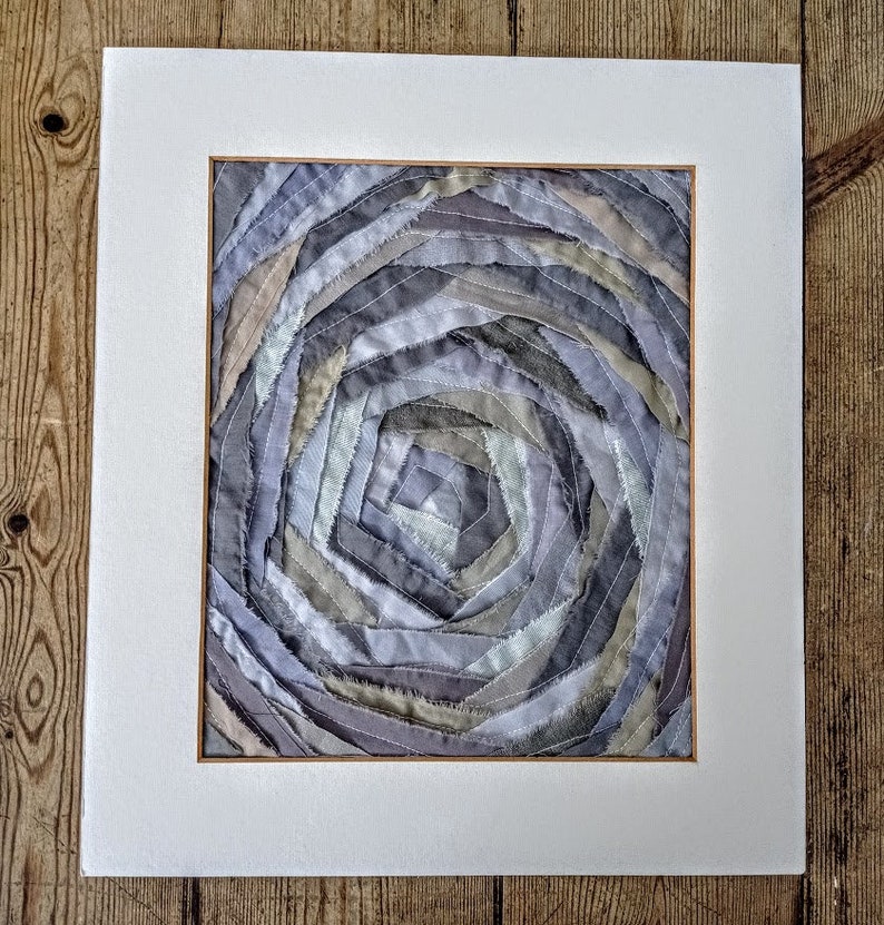 Pu&ograve; includere: Un'opera d'arte incorniciata che presenta un motivo a spirale di ritagli di tessuto in tonalit&agrave; di grigio, beige e bianco.