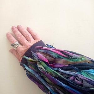 Puede incluir: Un primer plano de una camisa de manga larga azul oscuro con un ribete deshilachado multicolor. La camisa est&aacute; hecha de una tela suave y c&oacute;moda y tiene un estilo &uacute;nico y bohemio.