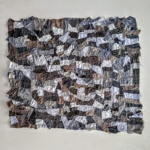 Op de afbeelding: Een patchworkdeken gemaakt van verschillende tinten grijs, bruin en wit. De stof is in onregelmatige vormen geknipt en aan elkaar genaaid om een ​​getextureerd oppervlak te cre&euml;ren.