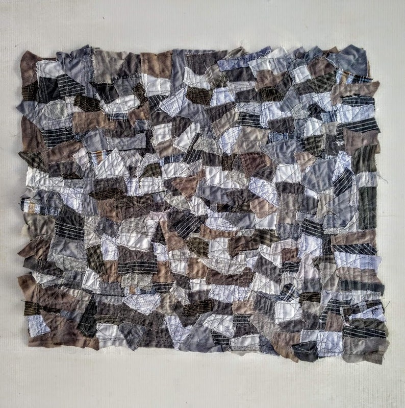 Op de afbeelding: Een patchworkdeken gemaakt van verschillende tinten grijs, bruin en wit. De stof is in onregelmatige vormen geknipt en aan elkaar genaaid om een ​​getextureerd oppervlak te cre&euml;ren.