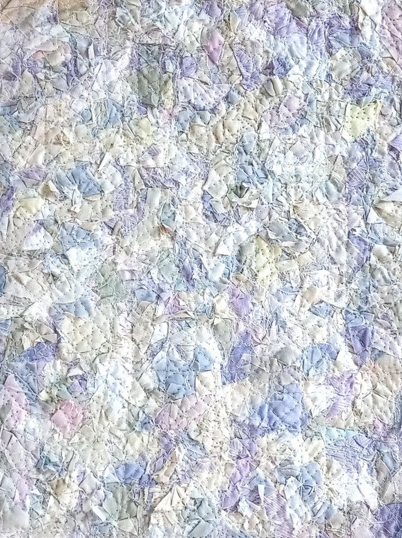 May include: A close-up of a quilt made from many small pieces of fabric in shades of white, blue, and purple. The fabric is sewn together in a patchwork pattern, creating a textured and intricate design.