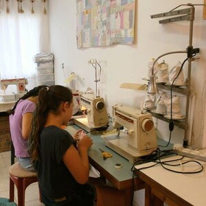 May include: Two people are sitting at a table with two sewing machines. The sewing machines are white and have a light blue table. The table has a light blue surface and a dark brown base. The people are wearing casual clothing. There is a colorful quilt hanging on the wall behind the table.