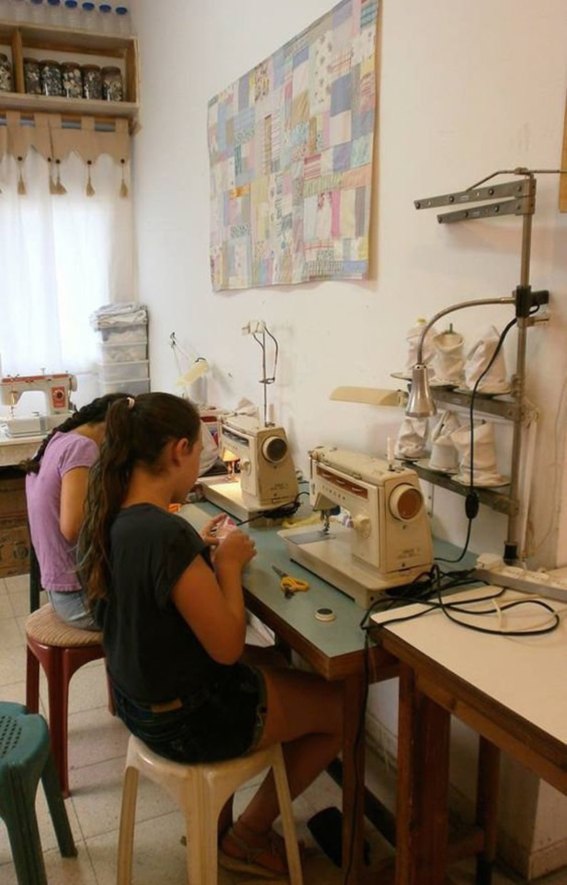 May include: Two people are sitting at a table with two sewing machines. The sewing machines are white and have a light blue table. The table has a light blue surface and a dark brown base. The people are wearing casual clothing. There is a colorful quilt hanging on the wall behind the table.