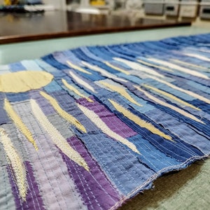 May include: A long, narrow wall hanging made of blue, purple and yellow fabric scraps. The fabric is sewn together in a patchwork style, creating a striped pattern. A large yellow circle is in the top left corner of the hanging.