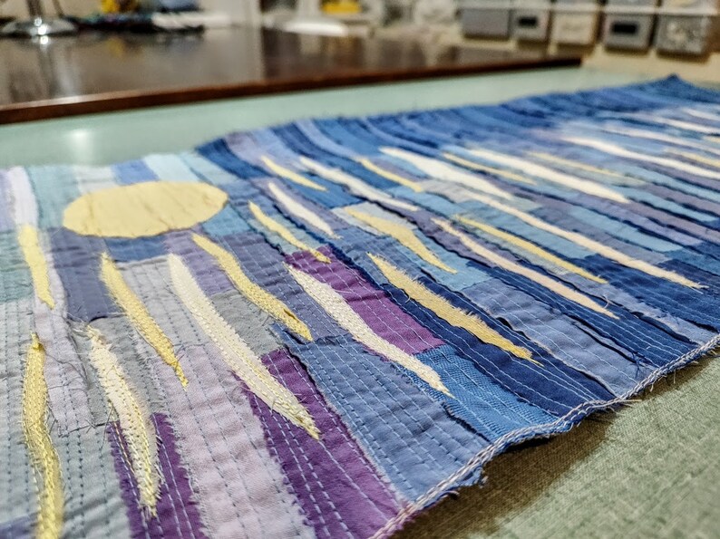 May include: A long, narrow wall hanging made of blue, purple and yellow fabric scraps. The fabric is sewn together in a patchwork style, creating a striped pattern. A large yellow circle is in the top left corner of the hanging.