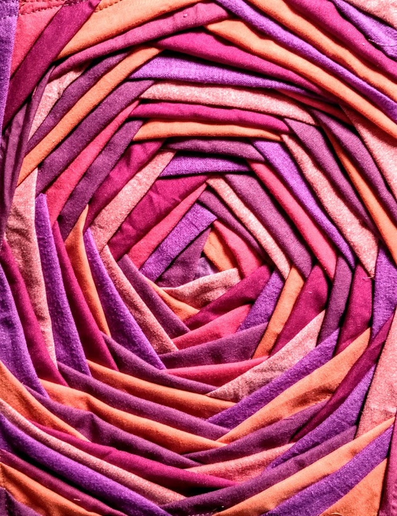 Pu&ograve; includere: Primo piano di un design circolare in tessuto realizzato con pi&ugrave; strisce di tessuto in tonalit&agrave; di rosa, viola e arancio. Il tessuto &egrave; intrecciato a spirale, creando una texture visivamente interessante.