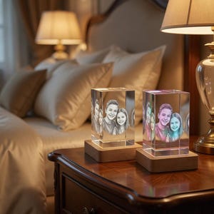 May include: Two illuminated crystal photo blocks with family portraits sit on a wooden nightstand. The blocks are rectangular and rest on wooden bases. A lamp with a beige shade is on the right, and a bed is in the background.