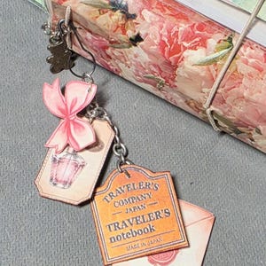 May include: A Traveler's Company notebook wrapped in floral paper, tied with twine, and adorned with charms. Charms include a pink bow, a perfume bottle, and a tag that reads "Traveler's Company Japan Traveler's Notebook Made in Japan."