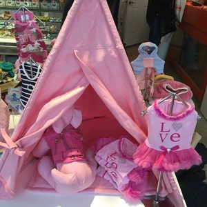 May include: A pink teepee tent with a pink stuffed animal inside. There are several pink and white outfits for small dogs hanging on hangers around the tent. One outfit has the word "Love" on it.