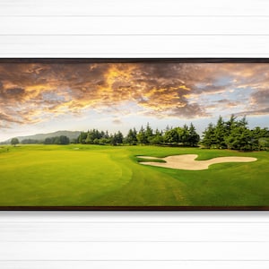 May include: A panoramic view of a golf course with a sand trap in the foreground. The course is surrounded by trees and a lake in the distance. The sky is a vibrant orange and pink, with fluffy white clouds.