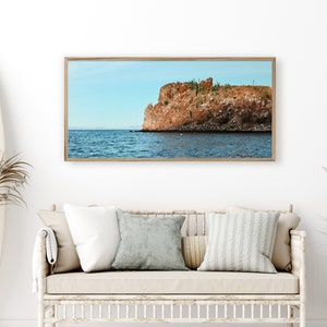 May include: A framed photograph of a rocky cliff with a blue sky and ocean in the background. The cliff is a reddish-brown color and has some green vegetation growing on it.