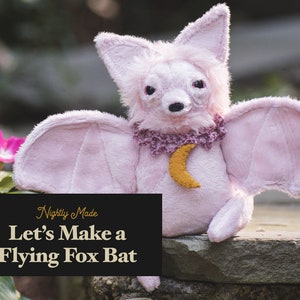 May include: A pink plush flying fox bat with a crescent moon charm. The bat is sitting on a rock with a green plant in the background. The text "Nightly Made Let's Make a Flying Fox Bat" is on a black background.