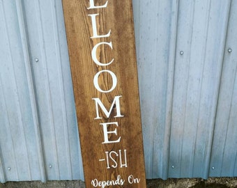 May include: A wooden sign with the word "WELCOME" written vertically in white paint. The sign also includes the text "ISH Depends On Who You Are & How Long You Stay!" with a leafy design at the top and bottom.